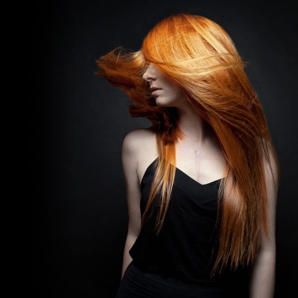 Porträt einer Frau mit langen, fließenden, roten Haaren, die sich in Bewegung befinden. Sie trägt ein schwarzes Top und posiert vor einem dunklen Hintergrund. Die professionelle Haarfarbe und das Styling wurden von Get Your Look in Frankfurt durchgeführt. Copyright by Get Your Look.